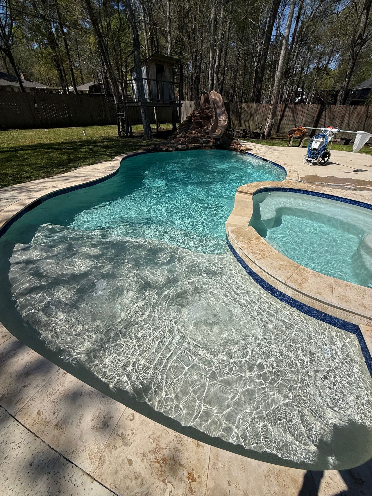 Freeform pool and attached spa with clear turquoise water, a slide and rock waterfall at the far end, and trees behind