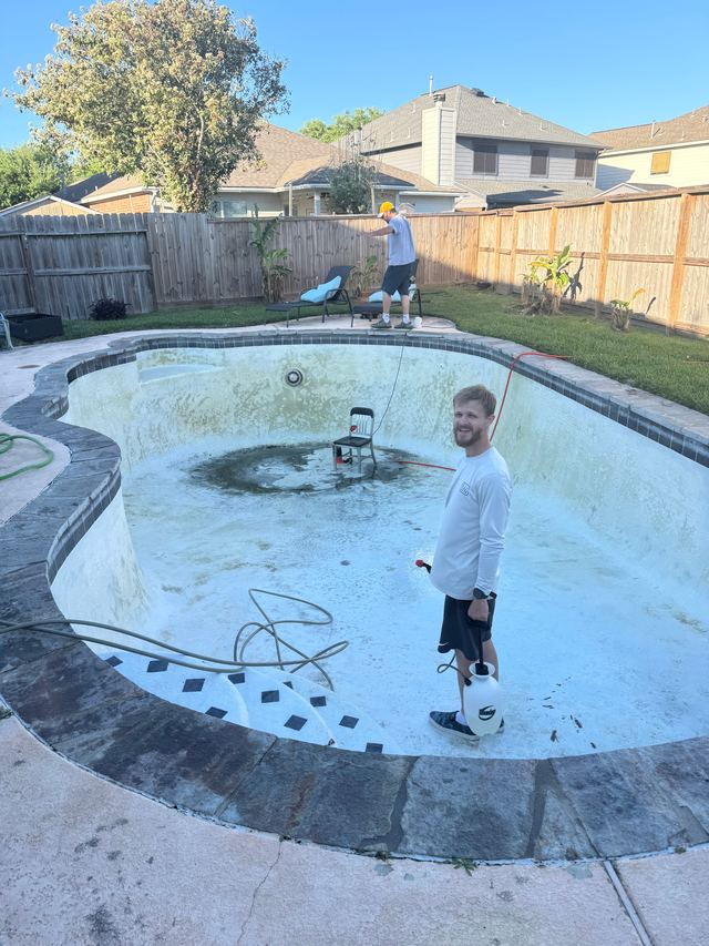 James Duncan smiling in a drained pool, holding a sprayer, with Aaron on the deck behind him